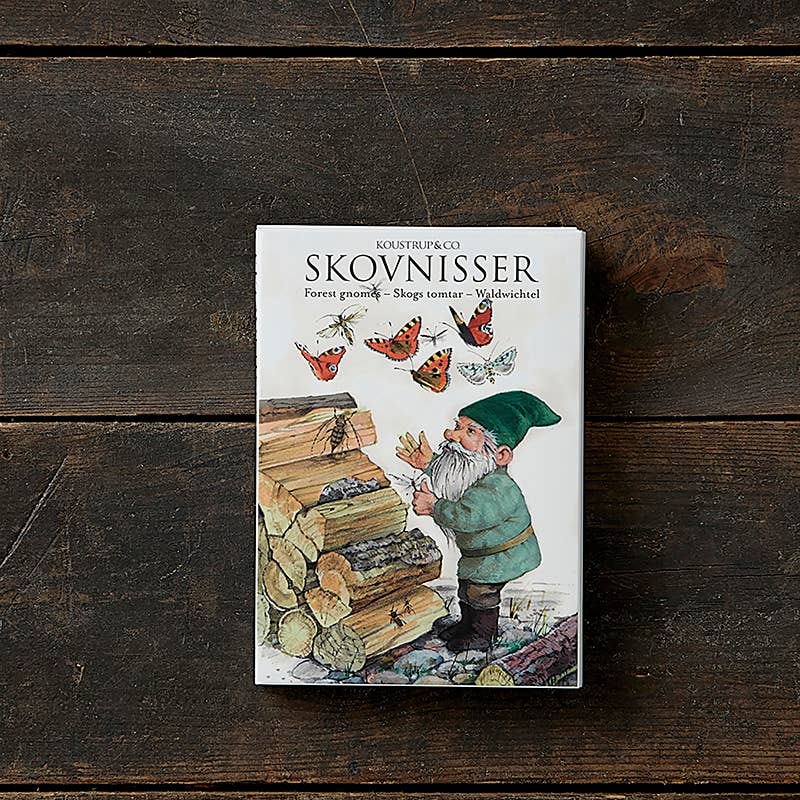 The cover of the card pack "Forrest Gnomes - Skovnisser," featuring eight different cards by Peter Nielsen Koustrupco, showcases a gnome stacking wood surrounded by fluttering butterflies. Set against a rustic wooden background, this captivating scene embodies the charm and whimsy typically linked to forest gnomes.