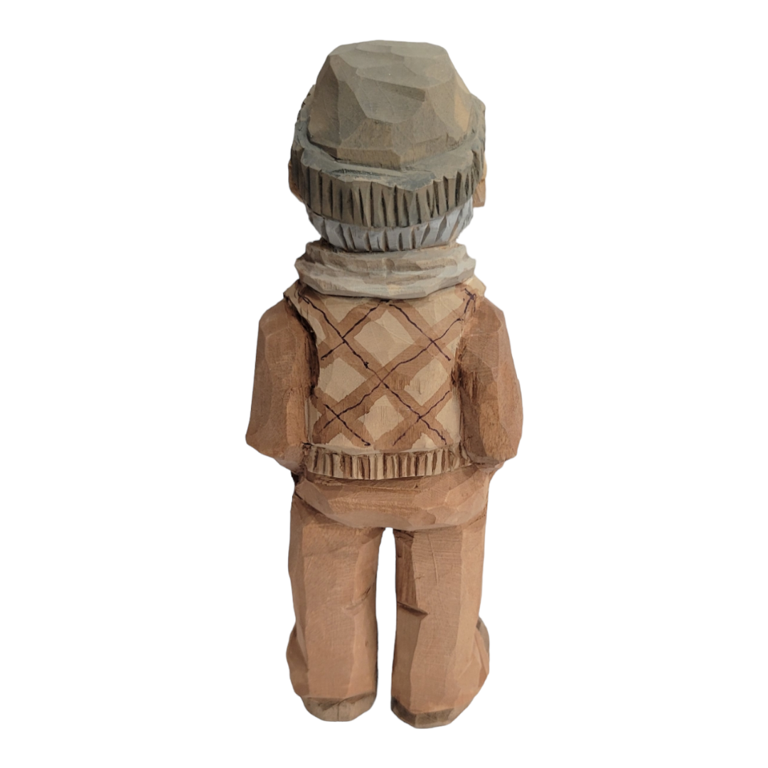 The 6 1/2 inch wood carved figurine, "Milo" by Bill Erickson, skillfully portrays a person wearing a hat and scarf, seen from the rear.