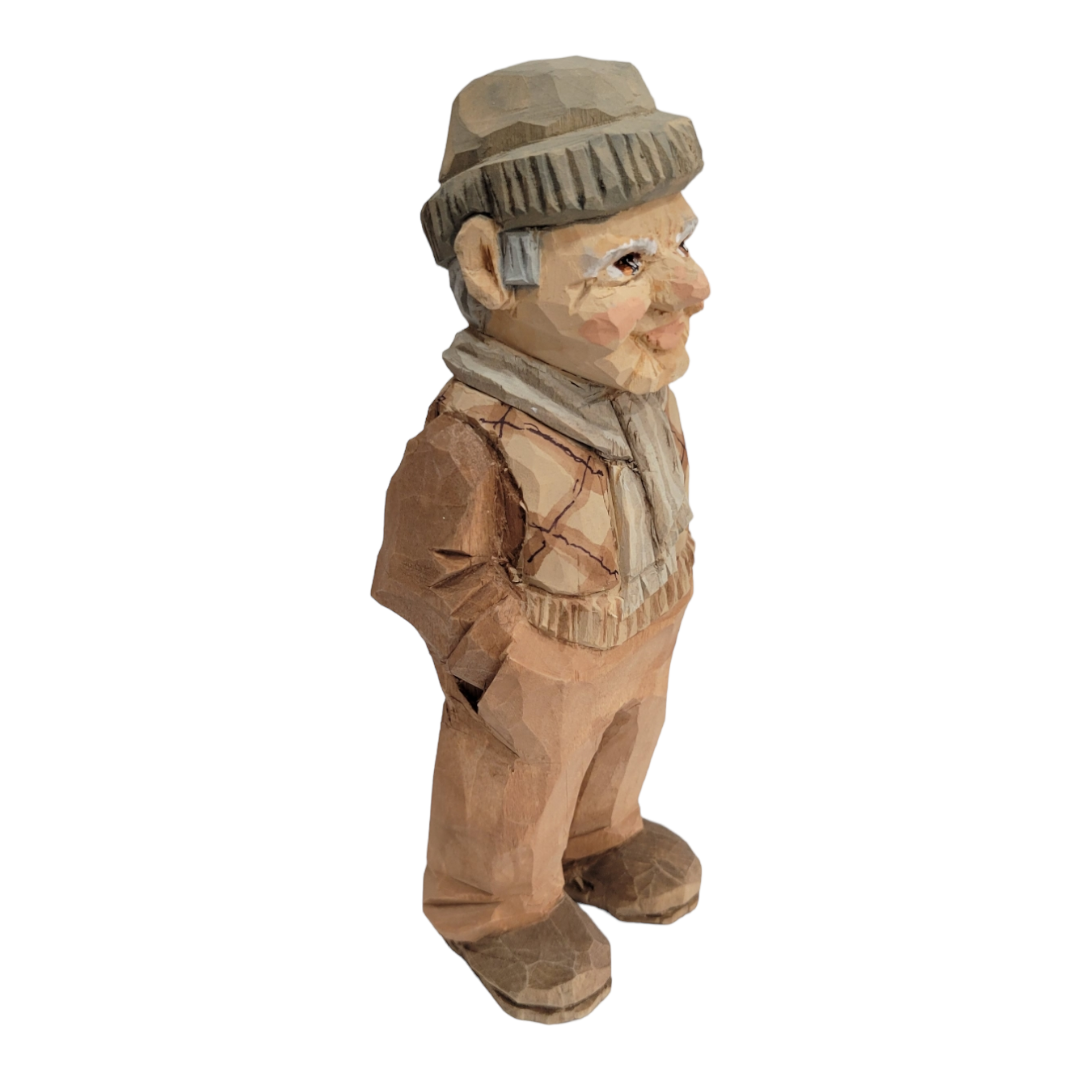 This 6 1/2 inch "Milo" wood carved figure by Bill Erickson portrays an elderly man clad in a cozy hat and scarf, standing with his hands in his pockets.
