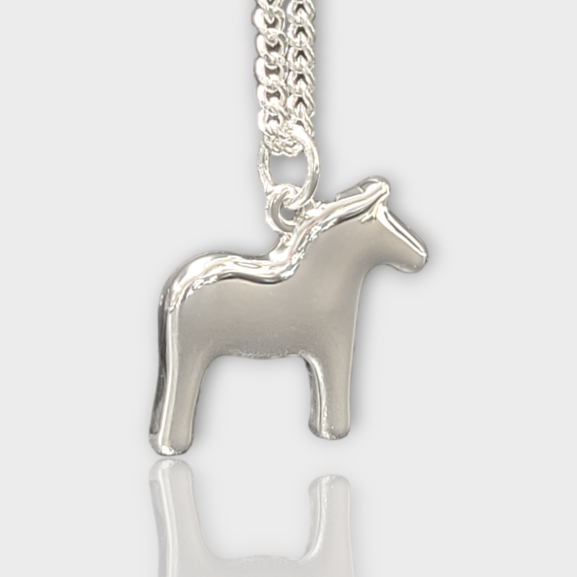 A silver pendant shaped like a horse, evocative of a classic Swedish emblem, hangs gracefully on a chain against a simple white backdrop. This Necklace: Dala Horse Necklace by Anna Viktoria beautifully showcases the allure of Scandinavian jewelry.