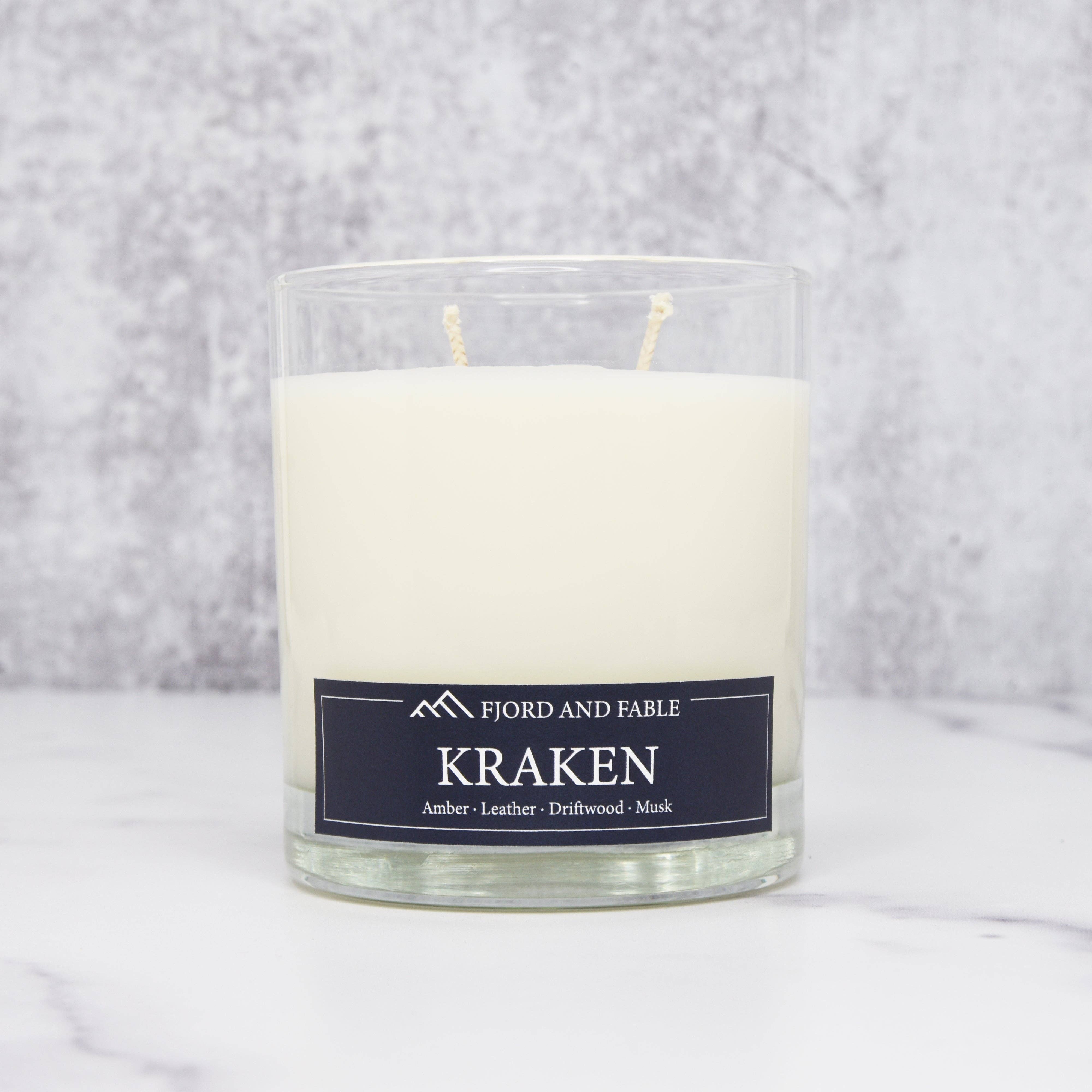 An 8oz Fjord & Fable Kraken candle in a clear glass jar, featuring amber, sea salt, driftwood, and leather notes, sits on a white surface with a gray background.