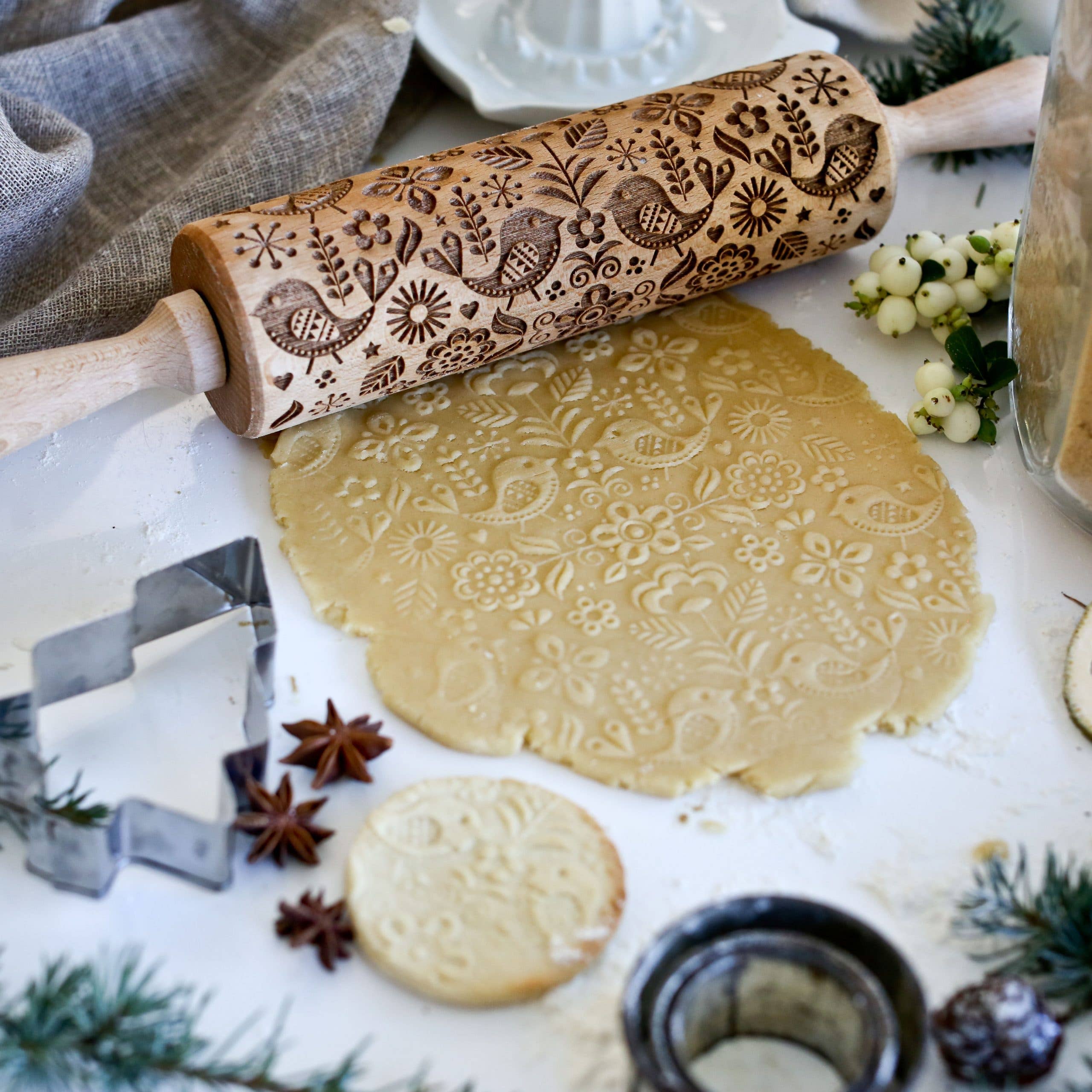 A Birds & Flowers Embossing Rolling Pin elegantly sits on intricately patterned cookie dough. Surrounding it are cookie cutters, spices, and greenery on a flour-dusted surface, capturing the essence of Scandinavian design—a delightful addition for crafting handmade gifts.