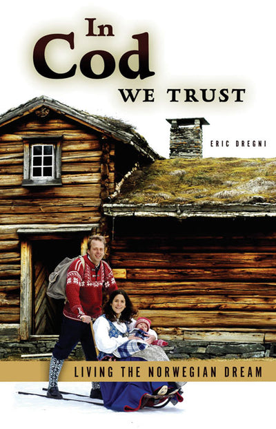 The cover of the book "In Cod We Trust" showcases a Norwegian-American family with a baby on a sled in front of a wooden cabin, capturing their journey depicted in the subtext, "Living the Norwegian Dream.