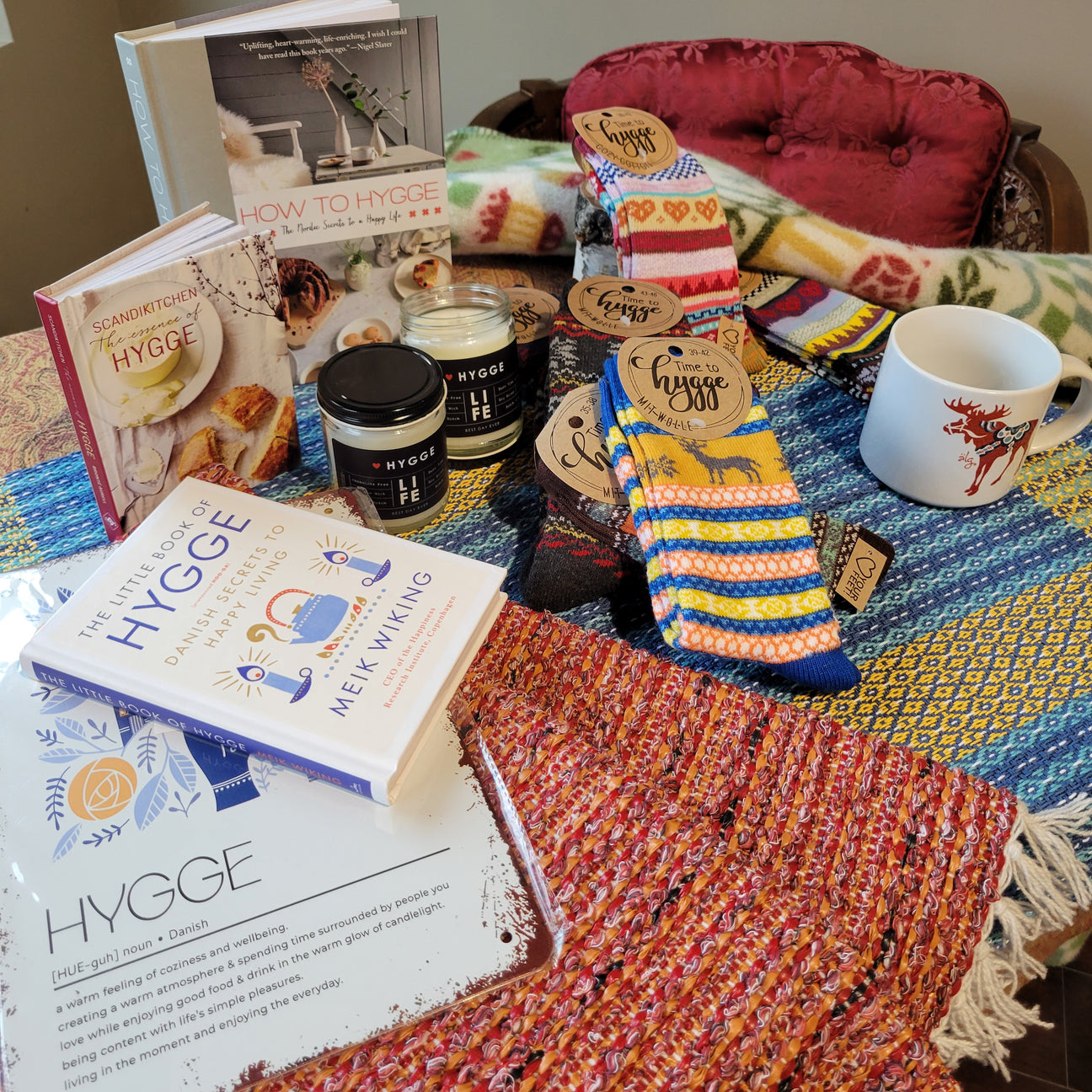 A cozy assortment of hygge-themed items, including books, socks, a candle, a mug, and a blanket, displayed on a chair.