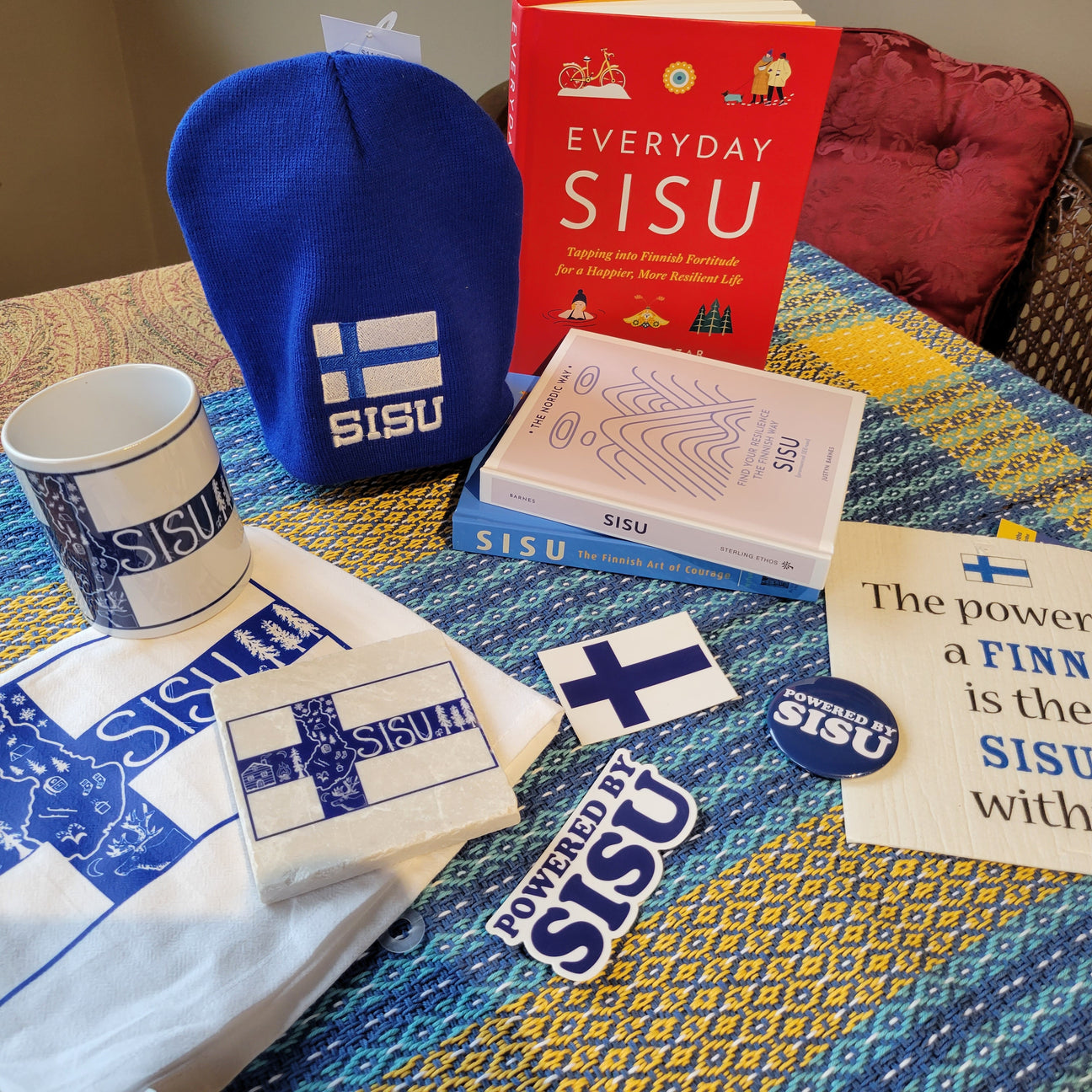 Collection of Sisu themed items, including a blue beanie, mug, books, tote bag, stickers, and a sign, displayed on a multicolored tablecloth.