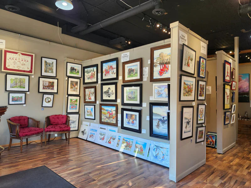 Art gallery interior with framed paintings and prints displayed on gray partition walls and several pieces resting on the floor; two red-cushioned chairs are in the corner on a wood floor.