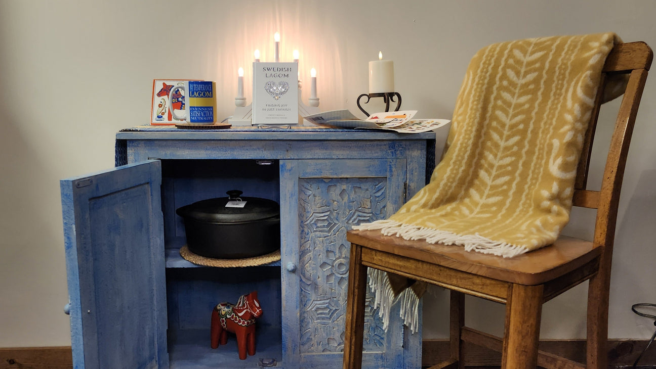 A wooden chair with a yellow patterned blanket next to a blue cabinet holding a black pot, decorative items, candles, and books on top.