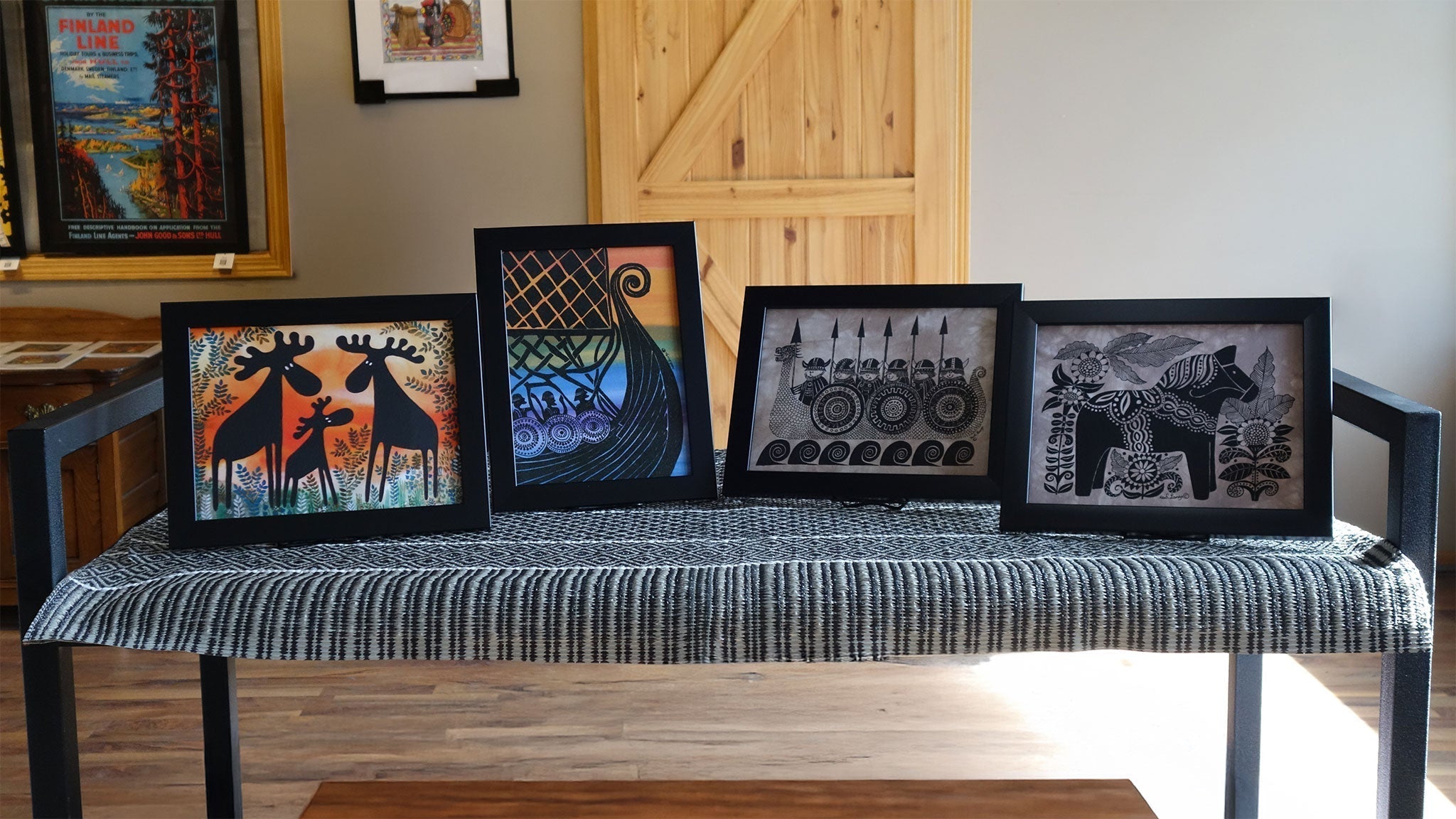 Four framed artworks on a table: two depict stylized animals, one has a colorful abstract design, and one shows a black and white geometric pattern.