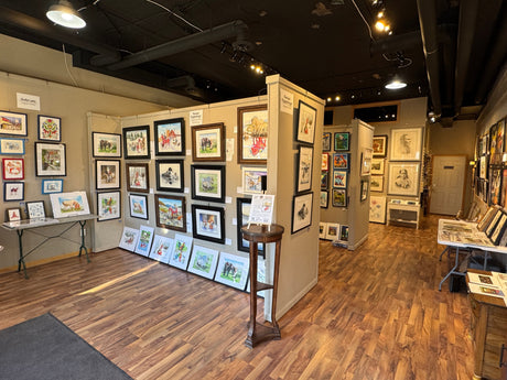 Art gallery interior featuring various framed paintings and drawings displayed on walls and panels, with a wooden floor and bright overhead lighting.