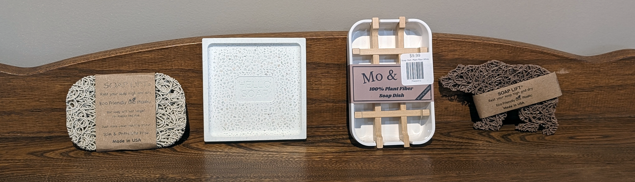 Four eco-friendly soap accessories on a wooden surface: a soap lift, a soap dish, a wood and plastic soap dish, and an exfoliating soap pouch.