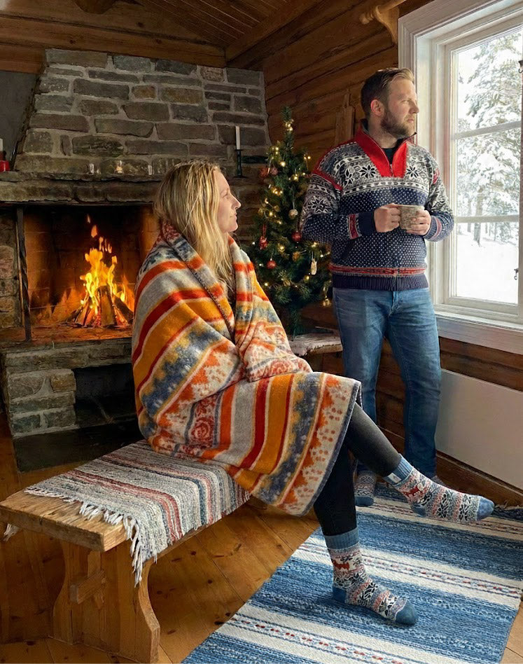 A display of colorful knit sweaters and woolen hats hangs on a wall in a store, featuring various patterns and designs. The floor is wooden and there are framed pictures above the display.