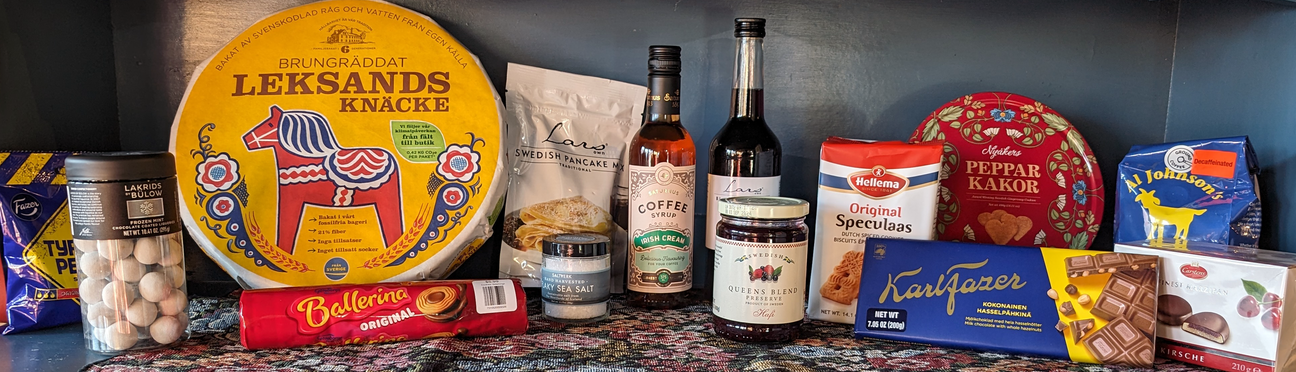 A variety of Swedish snacks, drinks, and treats are displayed on a patterned tablecloth, including Leksands knäckebröd, coffee, chocolate, and spiced cookies.