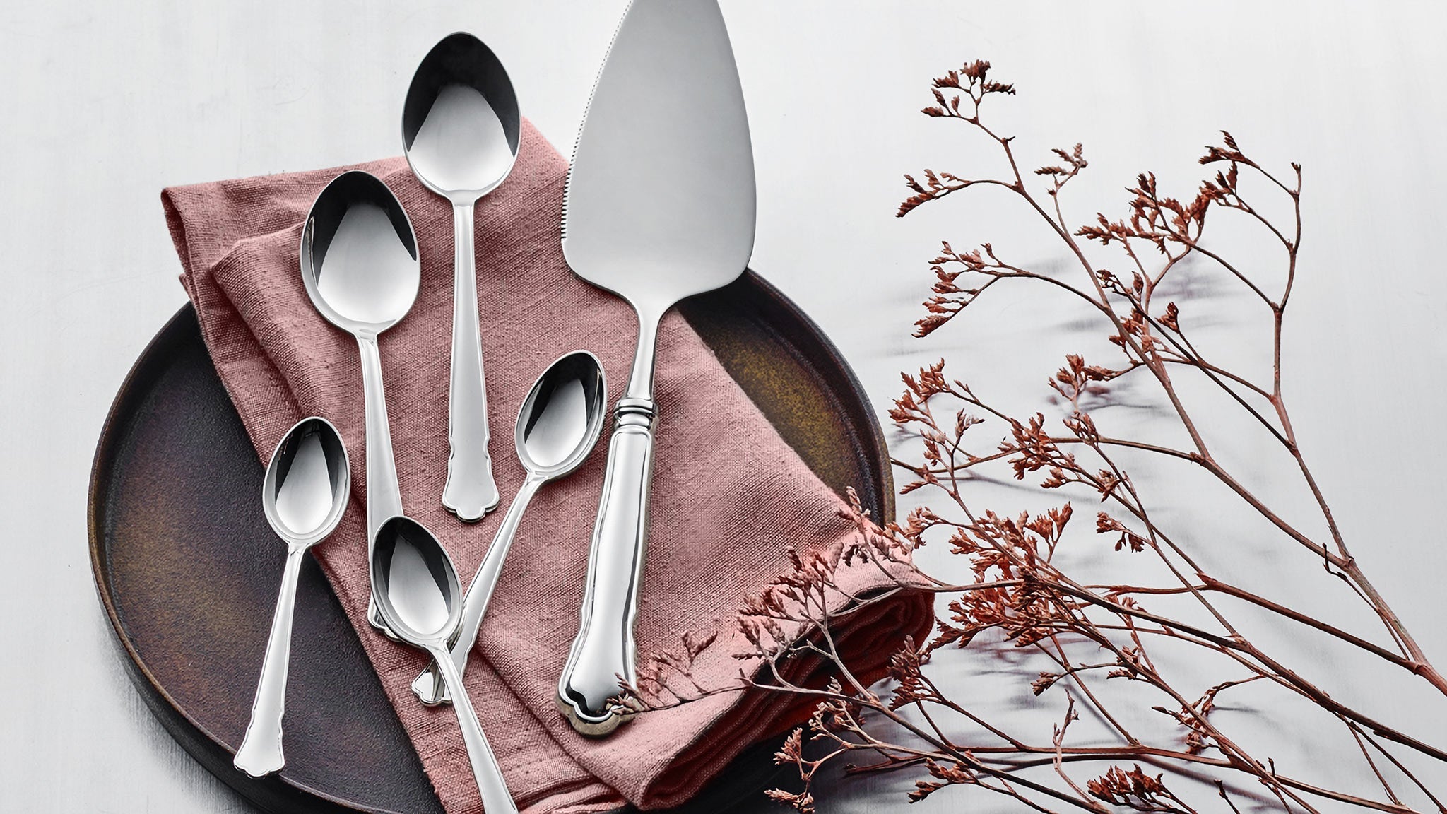 A plate and pink cloth with five silver utensils, including a cake server, are arranged beside dried branches on a light background.