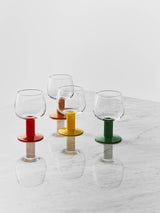 Four mouth-blown wine glasses with thick stems and circular bases in red, orange, yellow, and green, arranged on a marble surface.