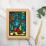 A greeting card titled "Evening at Camp Tent" by Sarah Angst features artwork of a red tent in a forest with orange leaves and a campfire, elegantly displayed on a marble surface alongside a pen and paperclips.