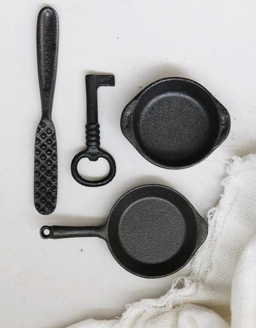 A variety of cast iron items, including a butter knife, key, two small pots, and a skillet sit on a light surface next to a white cloth—ideal for culinary tasks from everyday cooking to seafood preparation using the Skeppshult: Fish Scaler.