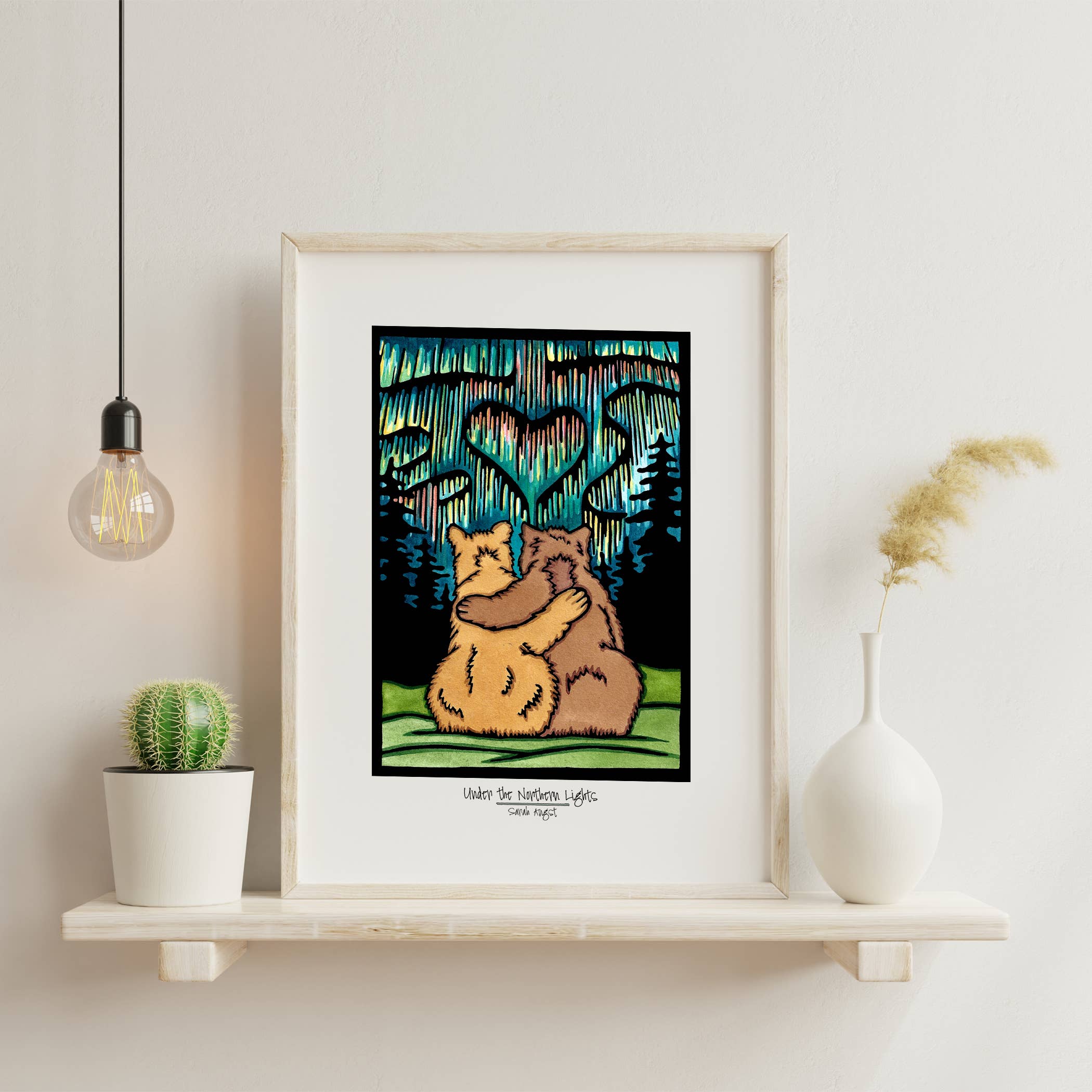 Framed 8x10 giclee print titled Under the Northern Lights by Sara Angst shows two bears hugging under heart-shaped lights, displayed on a shelf with a cactus, white vase, and hanging bulb.