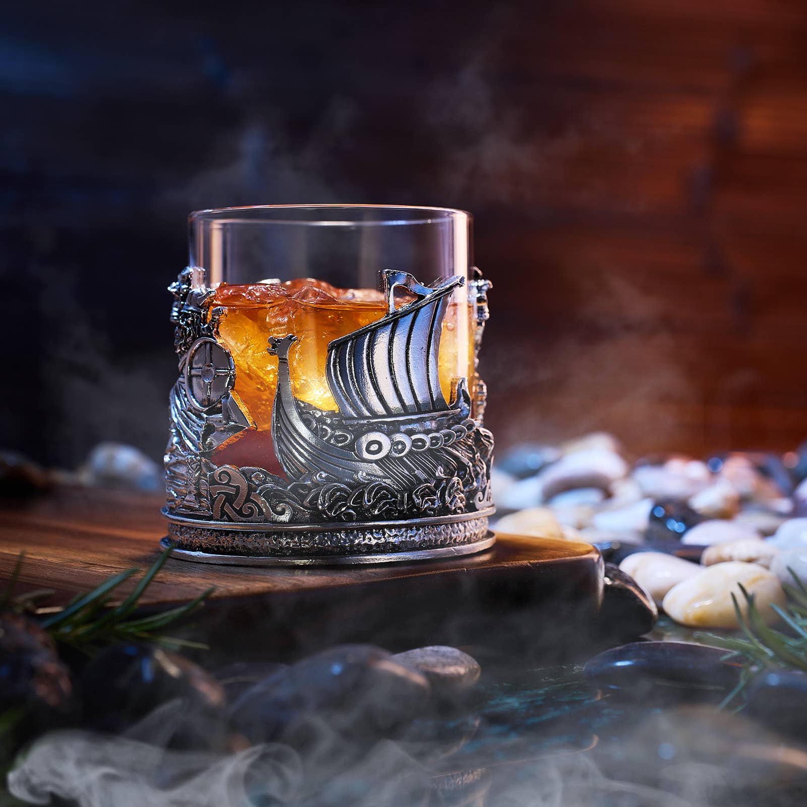 A glass of amber liquor with ice sits in a 12 oz Viking & Mythological Whiskey Drinking Glass, placed in a decorative metal holder on wood, with stones and mist in the foreground.