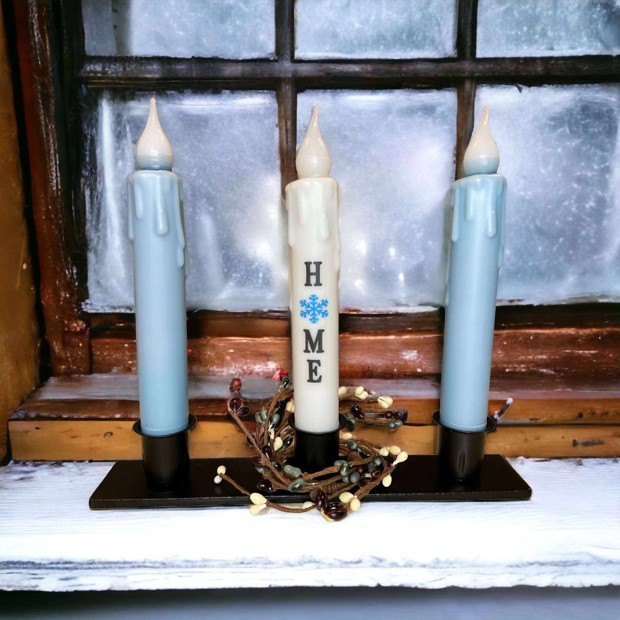 Three LED candles rest on a windowsill within a Triple Wrought Iron Taper Holder, with the center candle displaying the letters H M E alongside a snowflake symbol, forming a charming centerpiece. The frosted window suggests a cold atmosphere that complements the cozy candle designs.