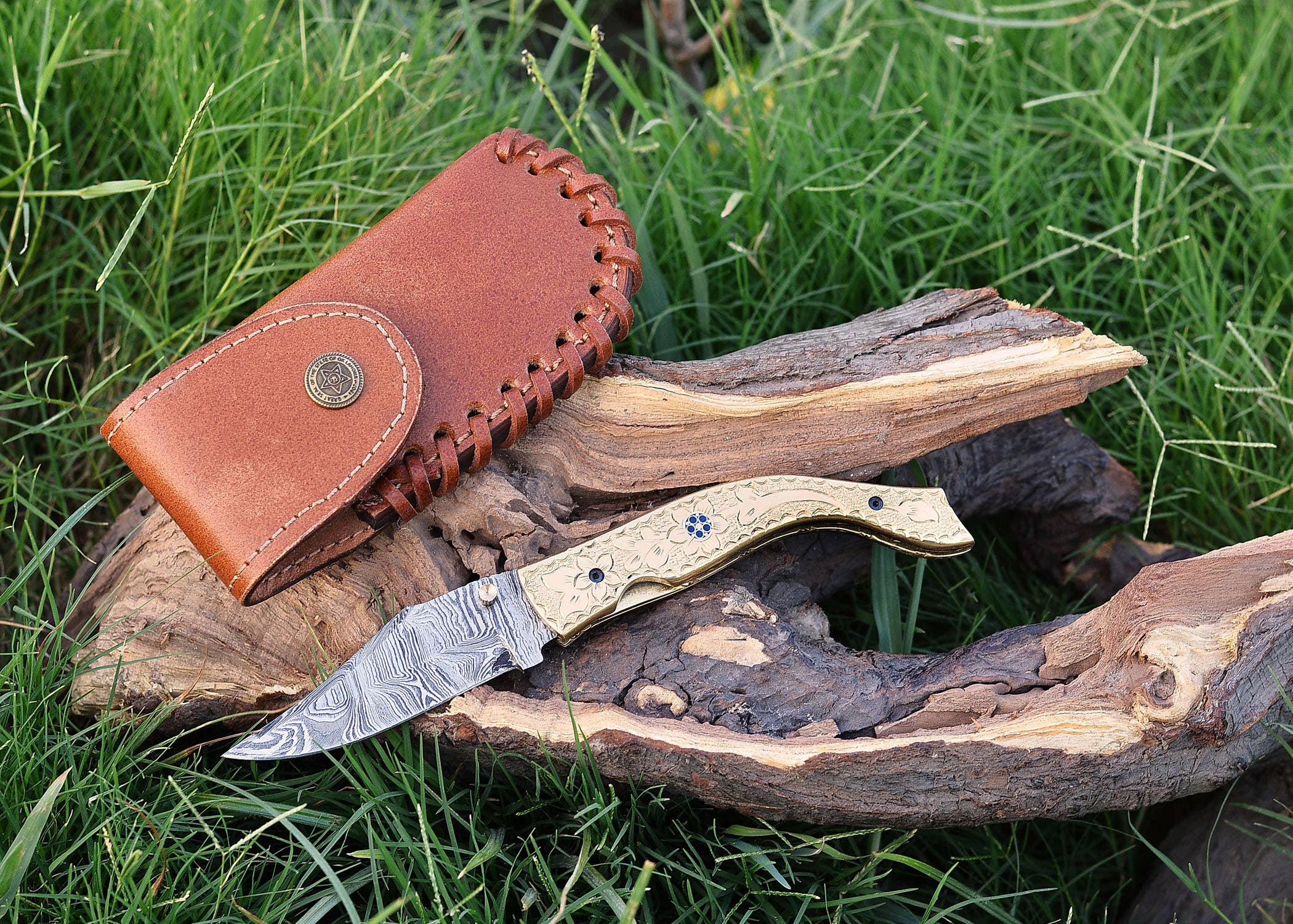 The Exquisite Golden Folding Knife, a luxury item with a stainless steel blade and intricate design, is displayed on wood in the grass. A brown leather sheath lies beside it.