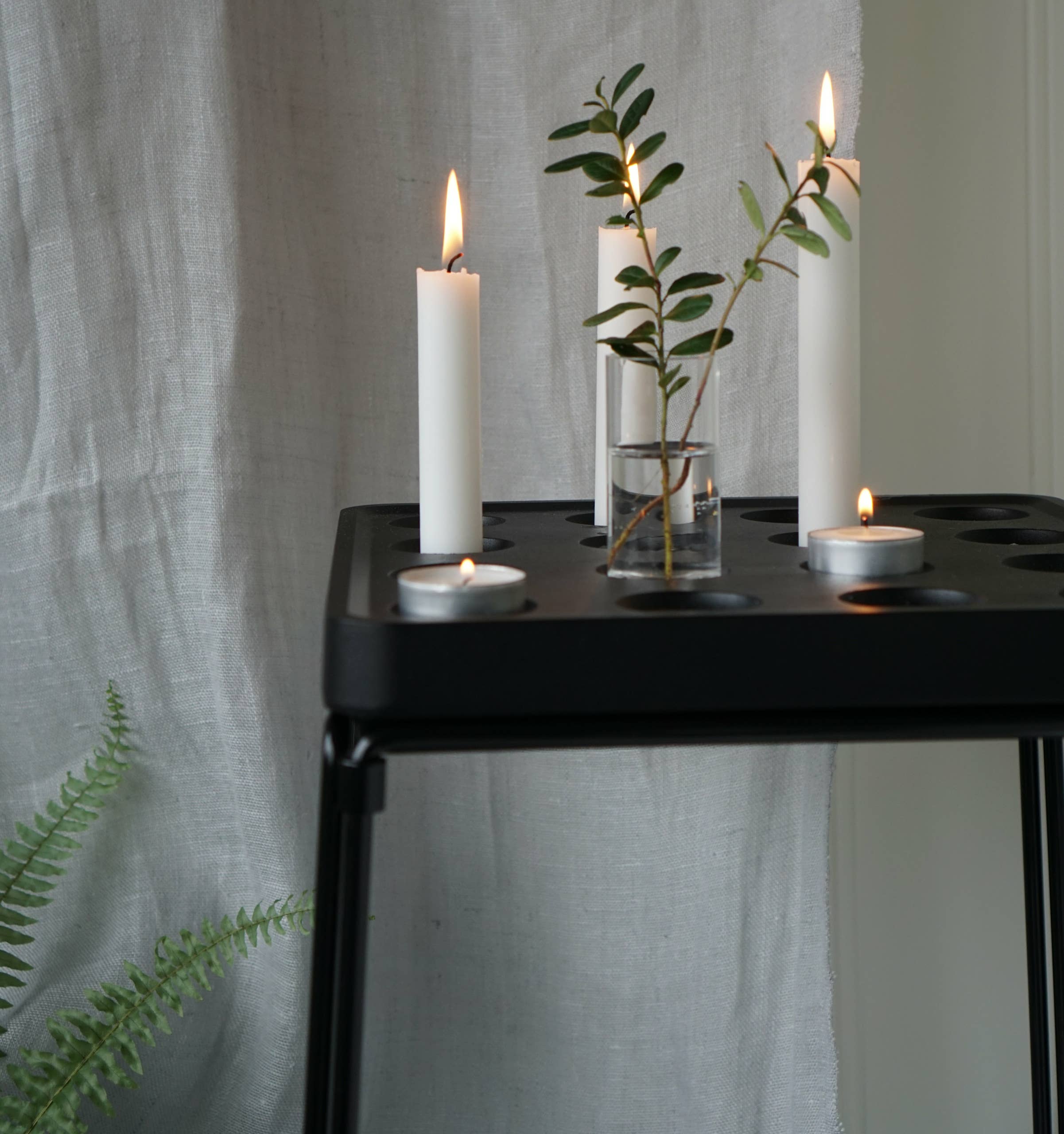 A black side table features a lit white taper candle in a Stumpastaken Candle Holder from the Mini Vases Pack of 4, alongside a small glass vase with green leaves. A light gray fabric backdrop and hints of a fern plant complete the setting.