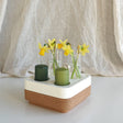 The Candle Holder: Mini Vases for Stumpastaken Candleholder 2-pack holds two green candles and three mini vases with yellow daffodils, beautifully arranged on a white and cork base against soft beige fabric for an elegant table display.