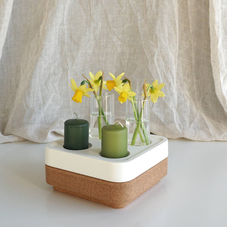 The Candle Holder: Mini Vases for Stumpastaken Candleholder 2-pack holds two green candles and three mini vases with yellow daffodils, beautifully arranged on a white and cork base against soft beige fabric for an elegant table display.