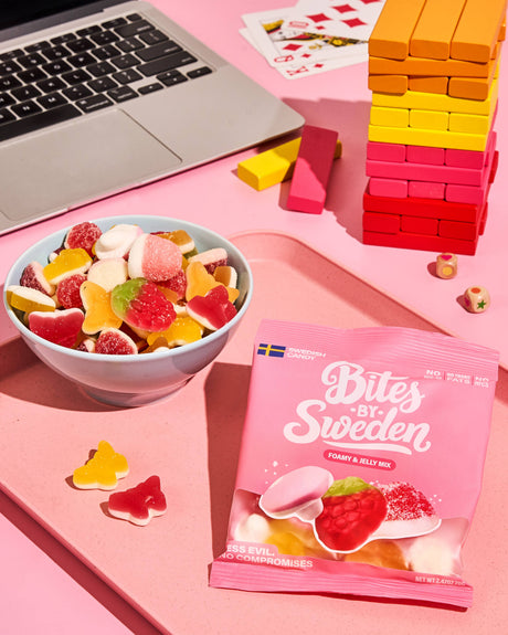 A bowl filled with assorted Swedish gummy candies next to a laptop, Jenga blocks, and a pink Bites by Sweden bag on a pink surface.