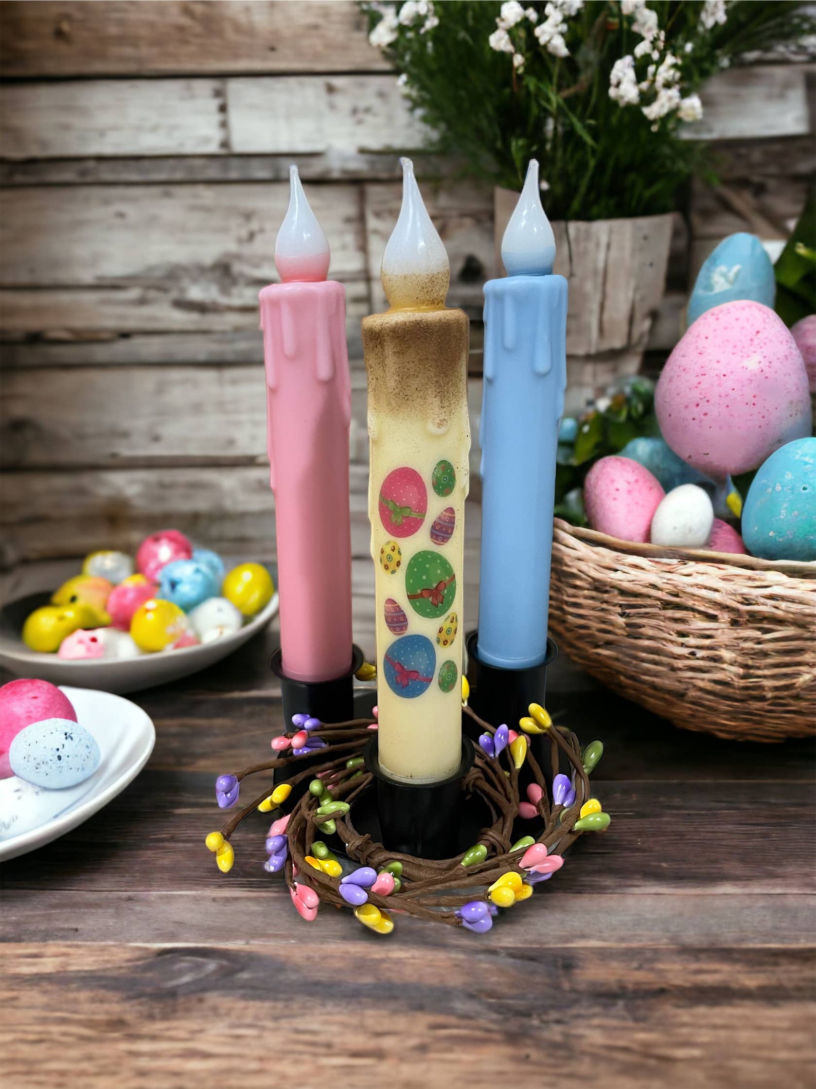 Three LED candles in pastel pink, cream, and blue are elegantly displayed on a Round Triple Wrought Iron Taper Holder atop a floral wreath. Colored eggs are arranged nearby in a basket and on a plate against a wooden backdrop adorned with delicate white flowers, capturing the timeless elegance of Lancaster blacksmith craftsmanship.