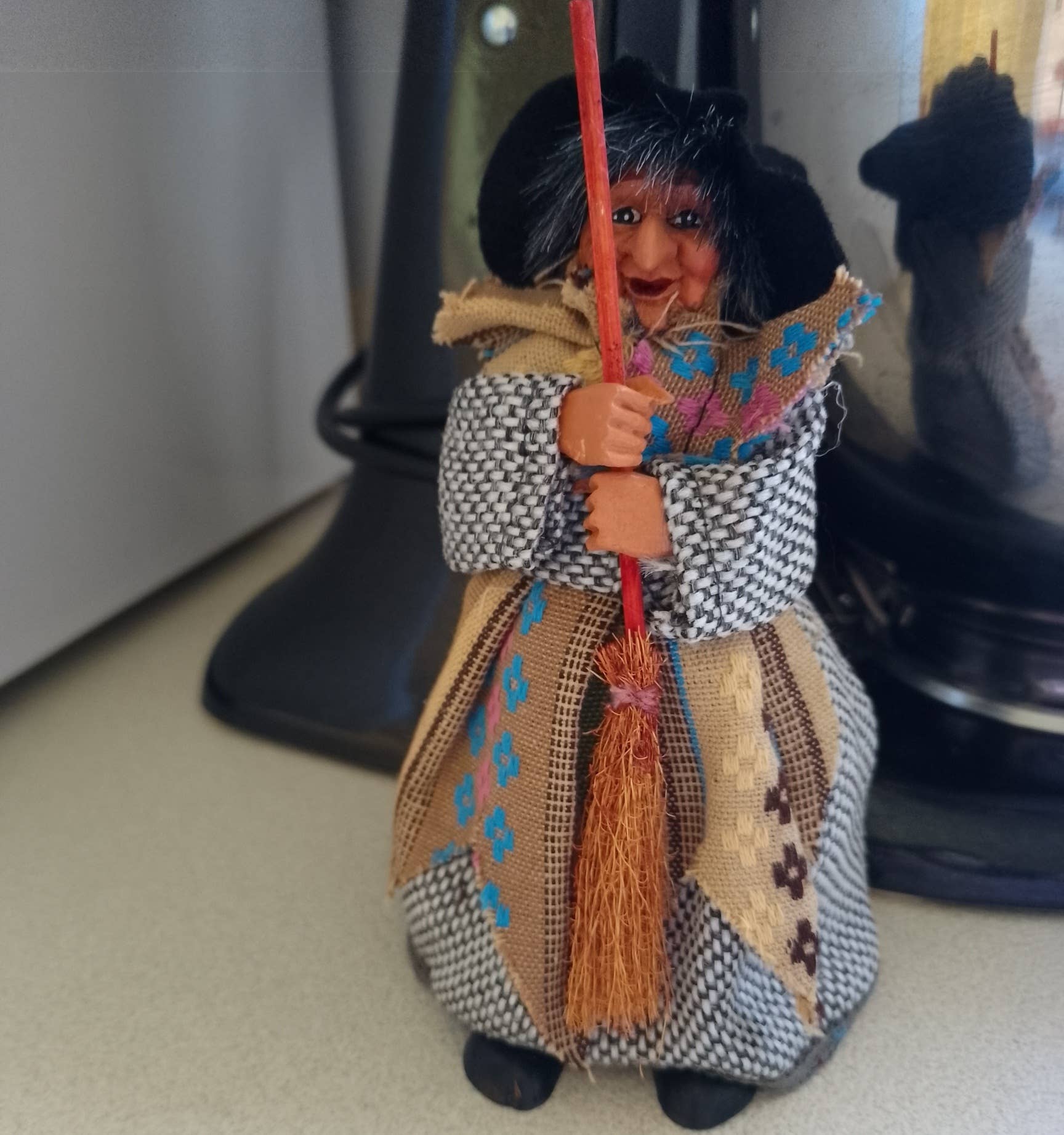The Kitchen Witch: Yasmina Beige-Gray stands 7" tall with black hair, a patterned dress, and a shawl, holding a broomstick—this charming doll is said to bring protection and good fortune to your kitchen.