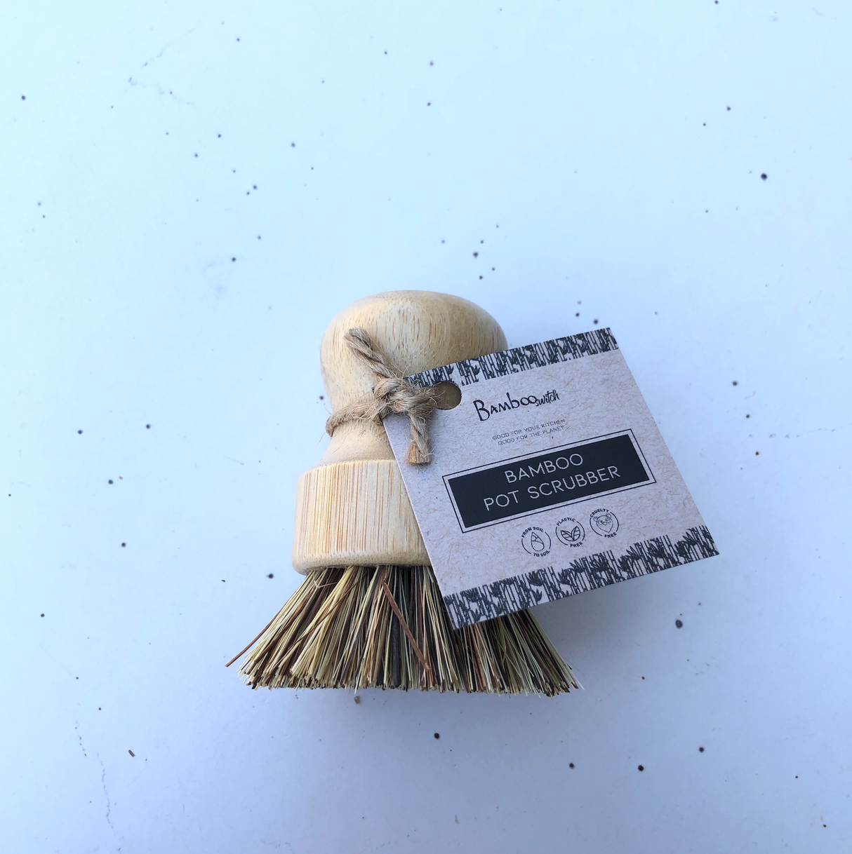 The Scrubber: Bamboo Pot Scruber features a wooden handle, natural bristles, and a brown paper tag—an eco-friendly, zero-waste kitchen alternative resting on a light surface.