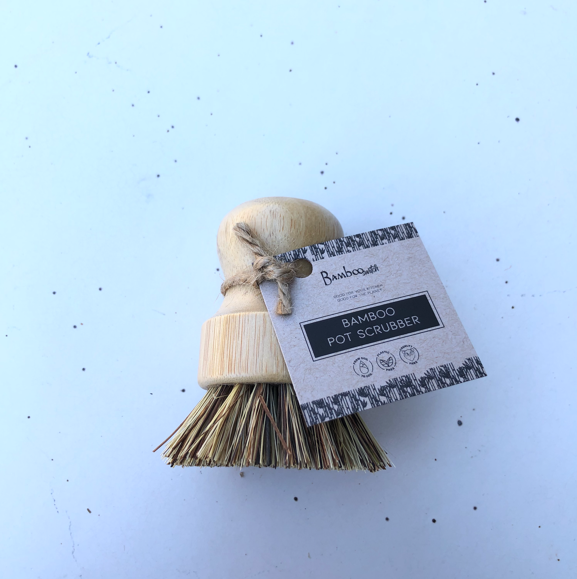The Scrubber: Bamboo Pot Scruber features a wooden handle, natural bristles, and a brown paper tag—an eco-friendly, zero-waste kitchen alternative resting on a light surface.