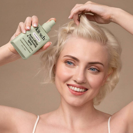 A smiling woman with short blonde hair holds Kitsch's Root Lifting & Thickening Spray (5 fl oz) near her head, ready to add volume and bounce—perfect for fuller-looking hair against a beige background.