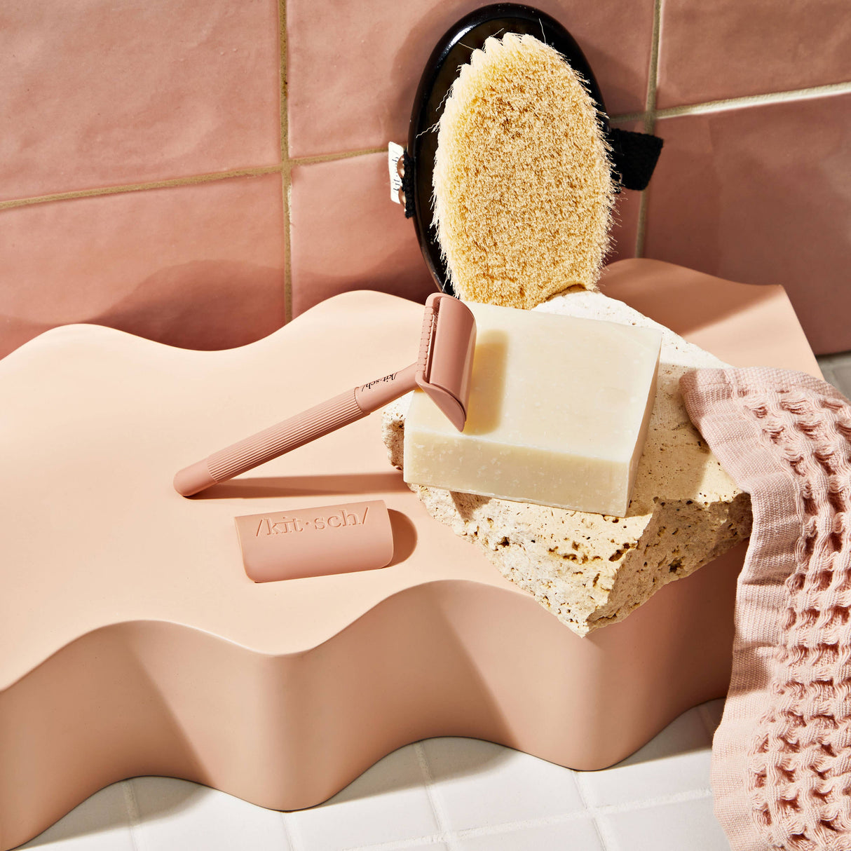 A pink razor, a sponge, a textured pink cloth, and KITSCH's Soap: Shave Butter Solid Bar 4 oz are arranged on a wavy, peach-colored surface against a tiled background—perfect companions for your favorite vegan or natural shaving routine.