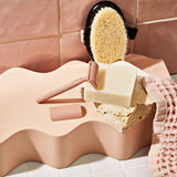 A pink razor, a sponge, a textured pink cloth, and KITSCH's Soap: Shave Butter Solid Bar 4 oz are arranged on a wavy, peach-colored surface against a tiled background—perfect companions for your favorite vegan or natural shaving routine.