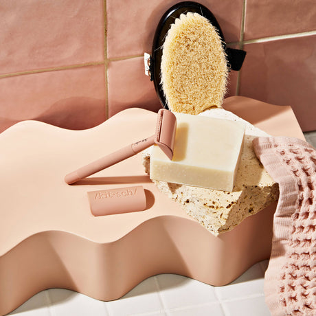 A pink razor, a sponge, a textured pink cloth, and KITSCH's Soap: Shave Butter Solid Bar 4 oz are arranged on a wavy, peach-colored surface against a tiled background—perfect companions for your favorite vegan or natural shaving routine.