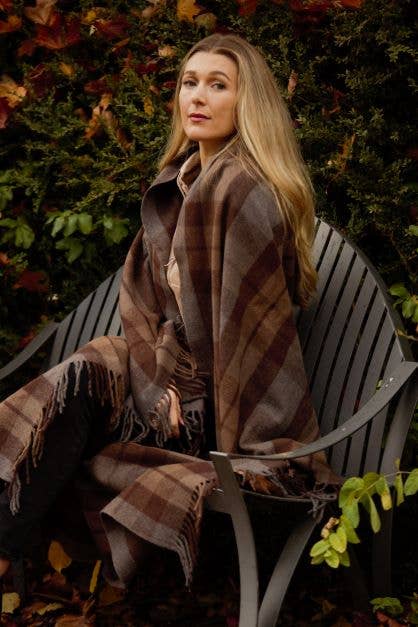 A woman with long blonde hair sits on a curved outdoor bench, wrapped in a cozy Outlander Merino Wool Blanket, surrounded by greenery and autumn leaves.