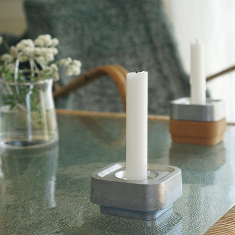A Stumpastaken Ettan candle holder in 100% recycled material with a white powder-coated candle (8.8x8.8x4 cm) sits on a glass table, while another candle and a vase of small white flowers accentuate the sustainable design.