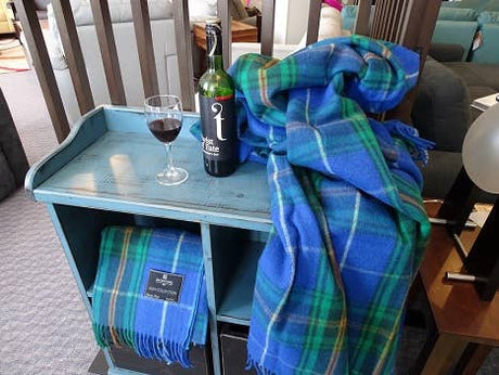 A glass and bottle of red wine sit on a small blue table next to a folded and draped Nova Scotia Deluxe Highland Tartan Blanket made of Merino Wool in blue and green hues.