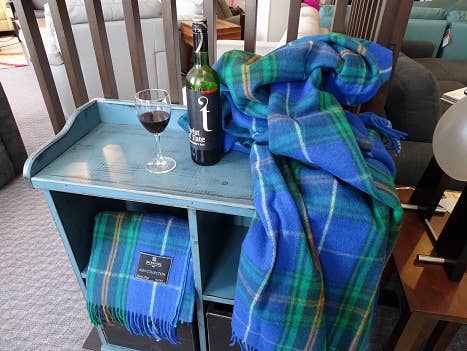 A glass and bottle of red wine sit on a small blue table next to a folded and draped Nova Scotia Deluxe Highland Tartan Blanket made of Merino Wool in blue and green hues.
