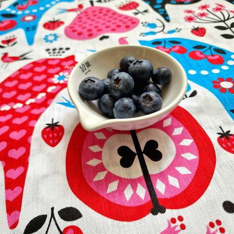 A small white measuring cup with blueberries rests on the "Tea Towel: Swedish Dala Horse Linen - Cotton Kitchen Towel" by Lotta Spjute, featuring hearts, flowers, fruit motifs, and a charming Swedish Dala Horse design.