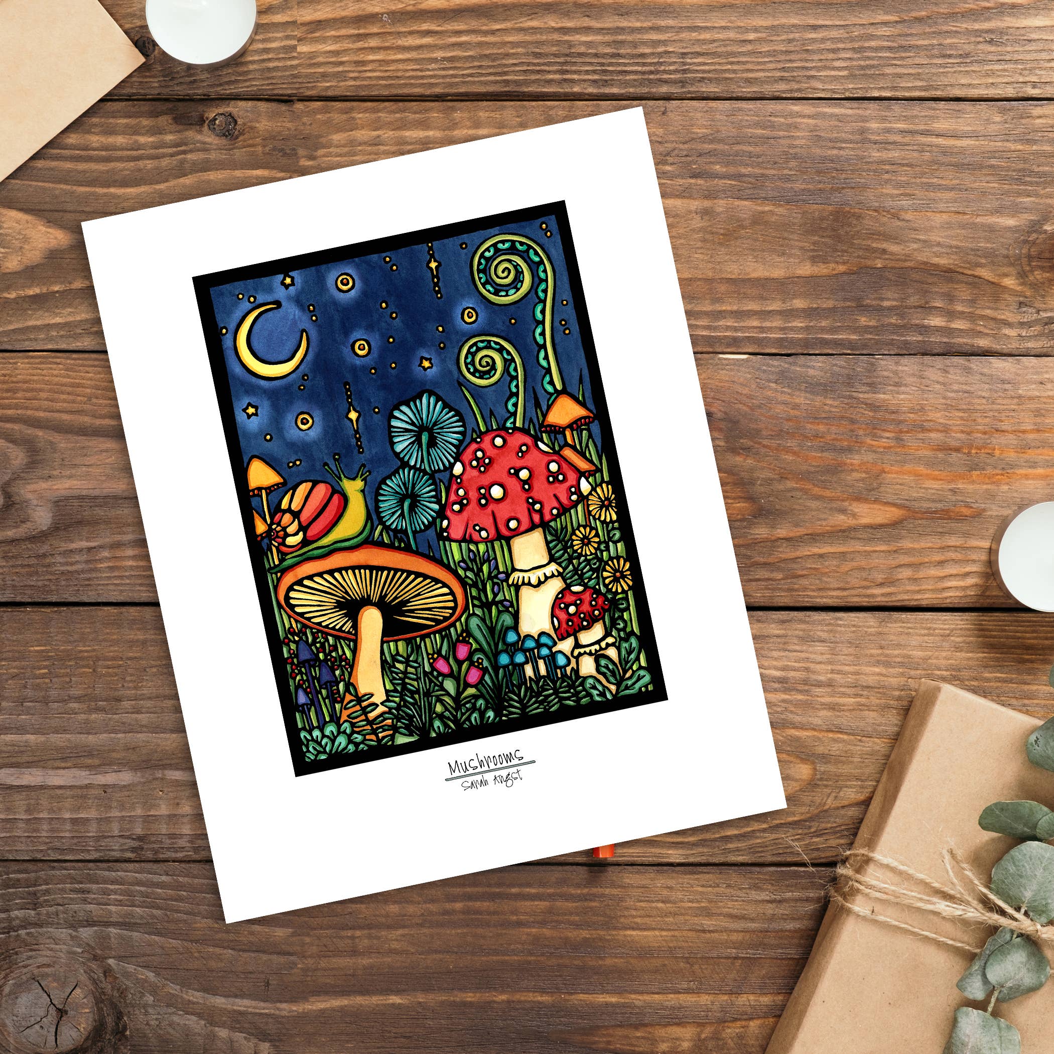 A vibrant giclee illustration titled "Artwork: Mushroom," measuring 11"x14" and elegantly framed in black, depicts mushrooms and plants under a starry sky on a wooden table, accompanied by a candle and a wrapped gift.