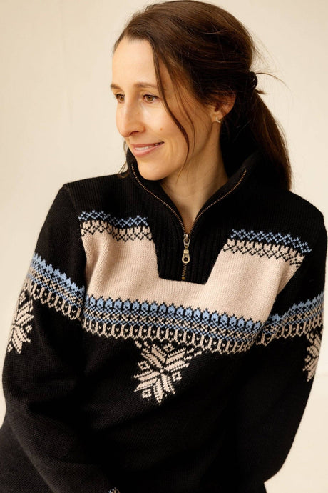 A woman wearing the Voss Women's Sweater (Black), made in Norway from 100% wool with blue and beige Nordic patterns, looks to the side and smiles slightly.