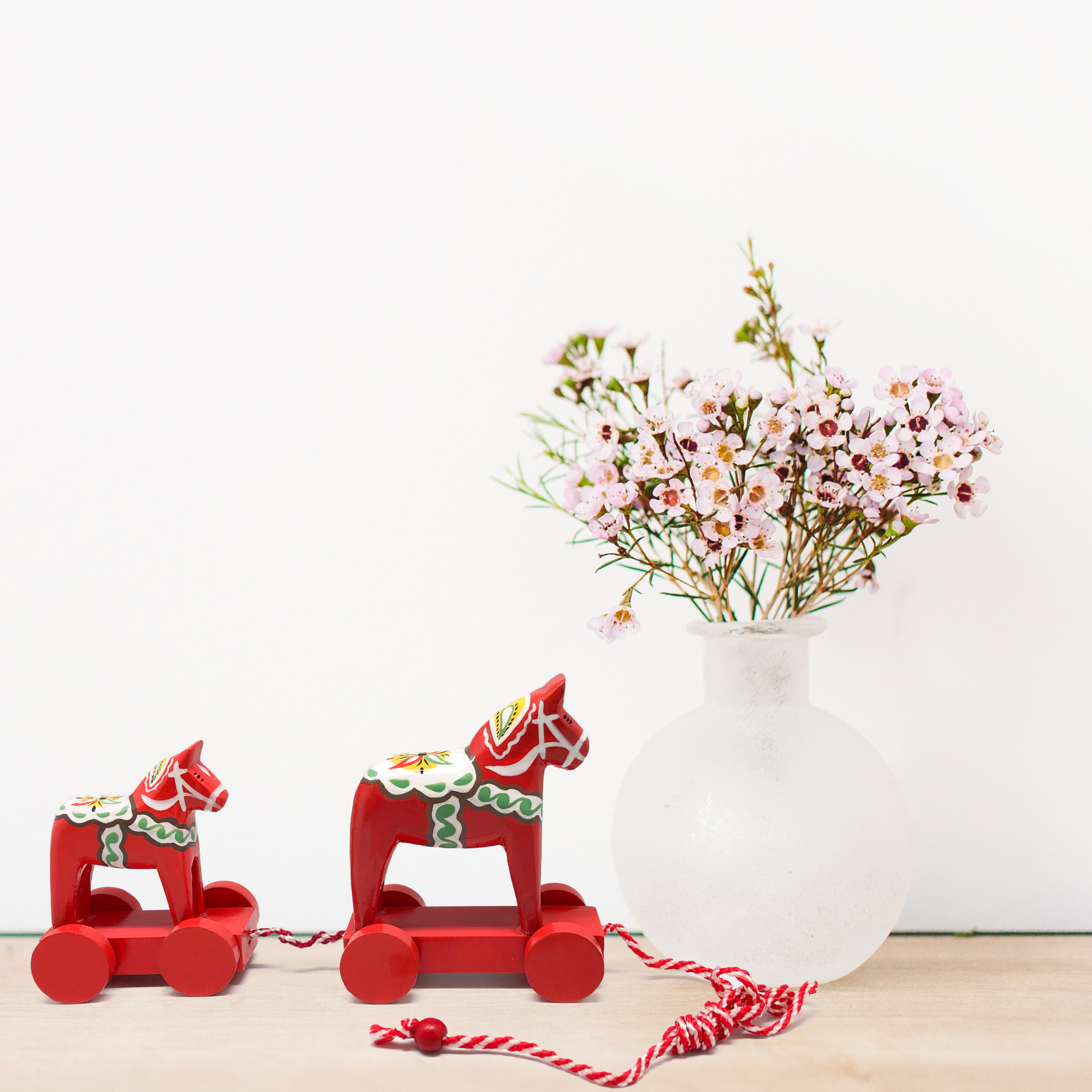 A Swedish-themed pull toy featuring two handcrafted red wooden Dala horses, adorned with decorative patterns and linked by a string, takes center stage. In the background, a white frosted vase filled with small pink flowers stands against a plain white wall, enhancing the Swedish decor aesthetic.