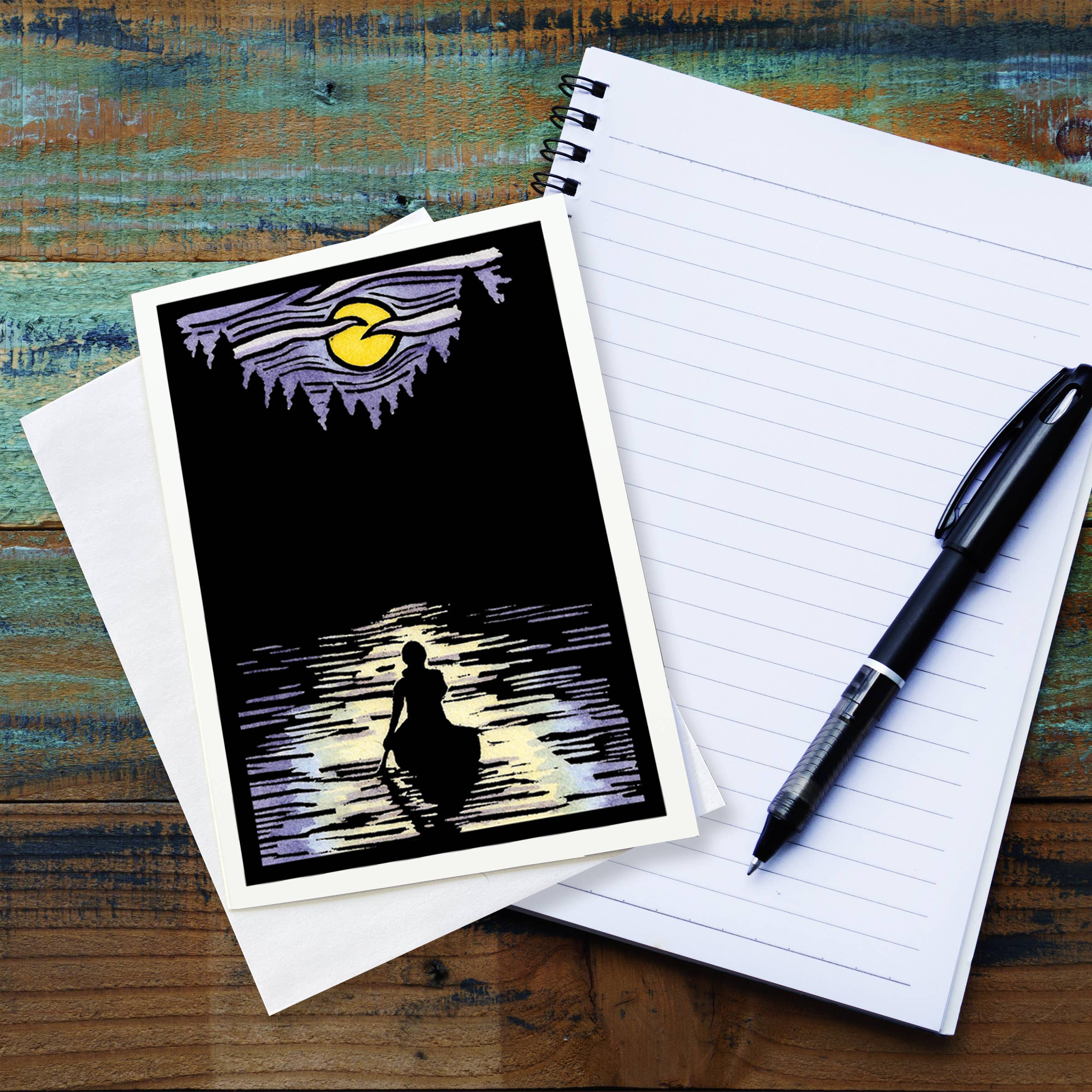 The "Solitude Canoe" greeting card by Sarah Angst, featuring a person in a boat silhouetted against a moonlit sky, is placed on top of a lined notebook next to a black pen on a wooden surface.