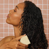A woman with wet, curly hair holds Kitsch's Coconut Oil Shampoo & Conditioner Bar Combo for Dry Damaged Hair (2x2.7oz) against her strands in front of a pink tiled wall.