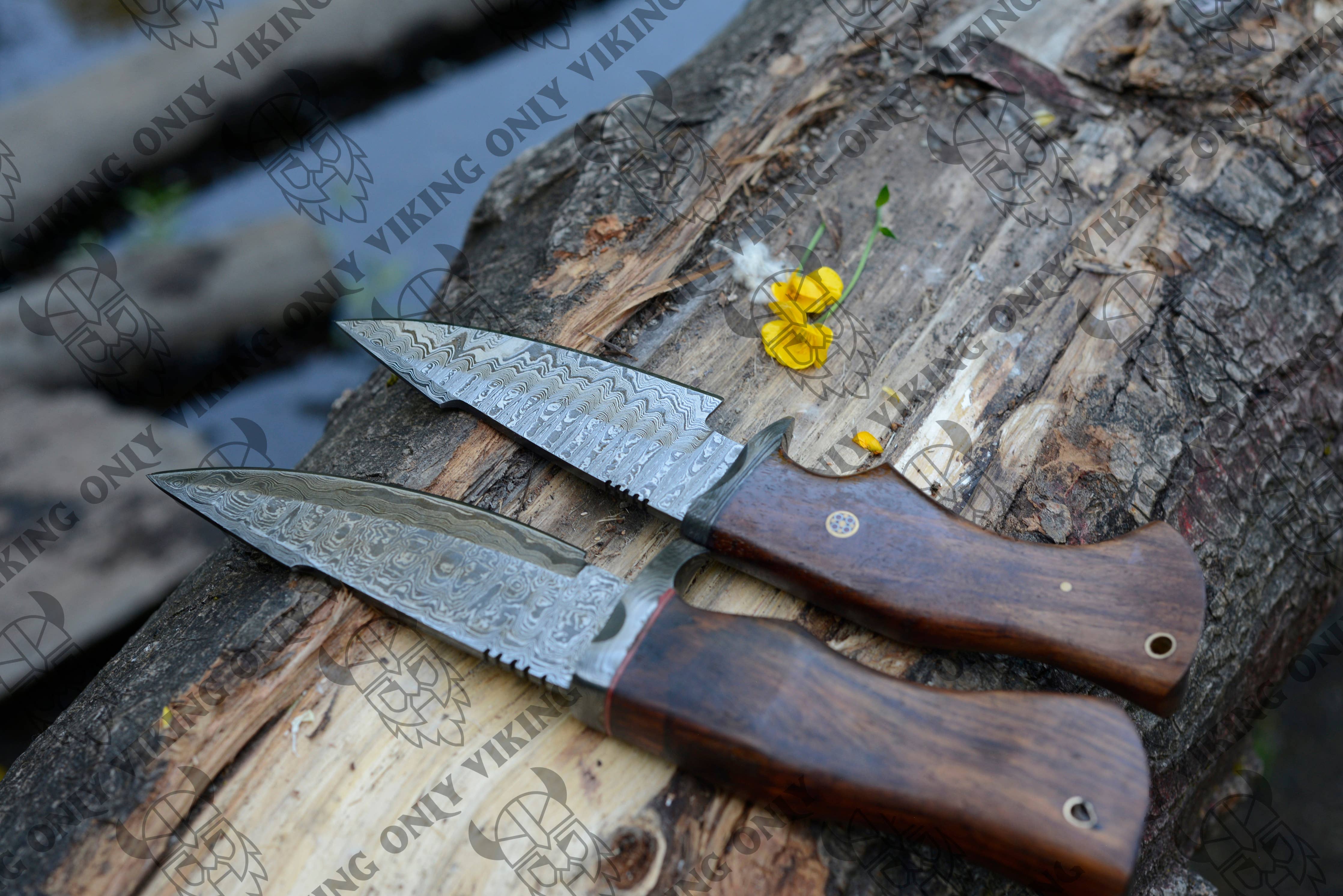 Two Premium Quality Damascus Knives with wooden handles rest on a log, their blades exhibiting the distinctive and intricate Damascus pattern. A yellow flower lies nearby, lending a touch of nature to this elegant display of camping tools.