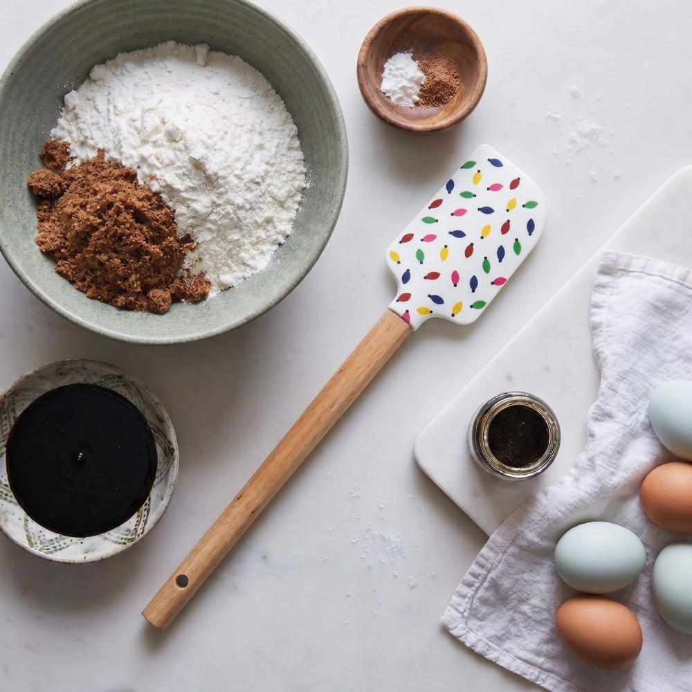 A spatula decorated with Christmas lights lies on a table surrounded by bowls of flour, brown sugar, eggs, molasses, and spices.