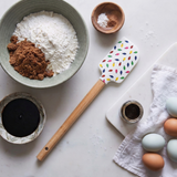 A spatula decorated with Christmas lights lies on a table surrounded by bowls of flour, brown sugar, eggs, molasses, and spices.