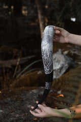 Someone holds the Horn: Raven Viking Drinking Horn w/ Metal Stand (14"-18"), featuring engraved patterns and a raven motif, outdoors near rocks and foliage.